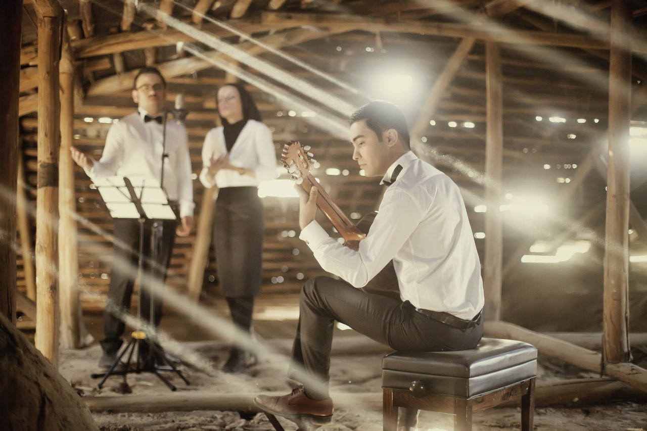 A seated guitarist and two standing performers in a wooden interior