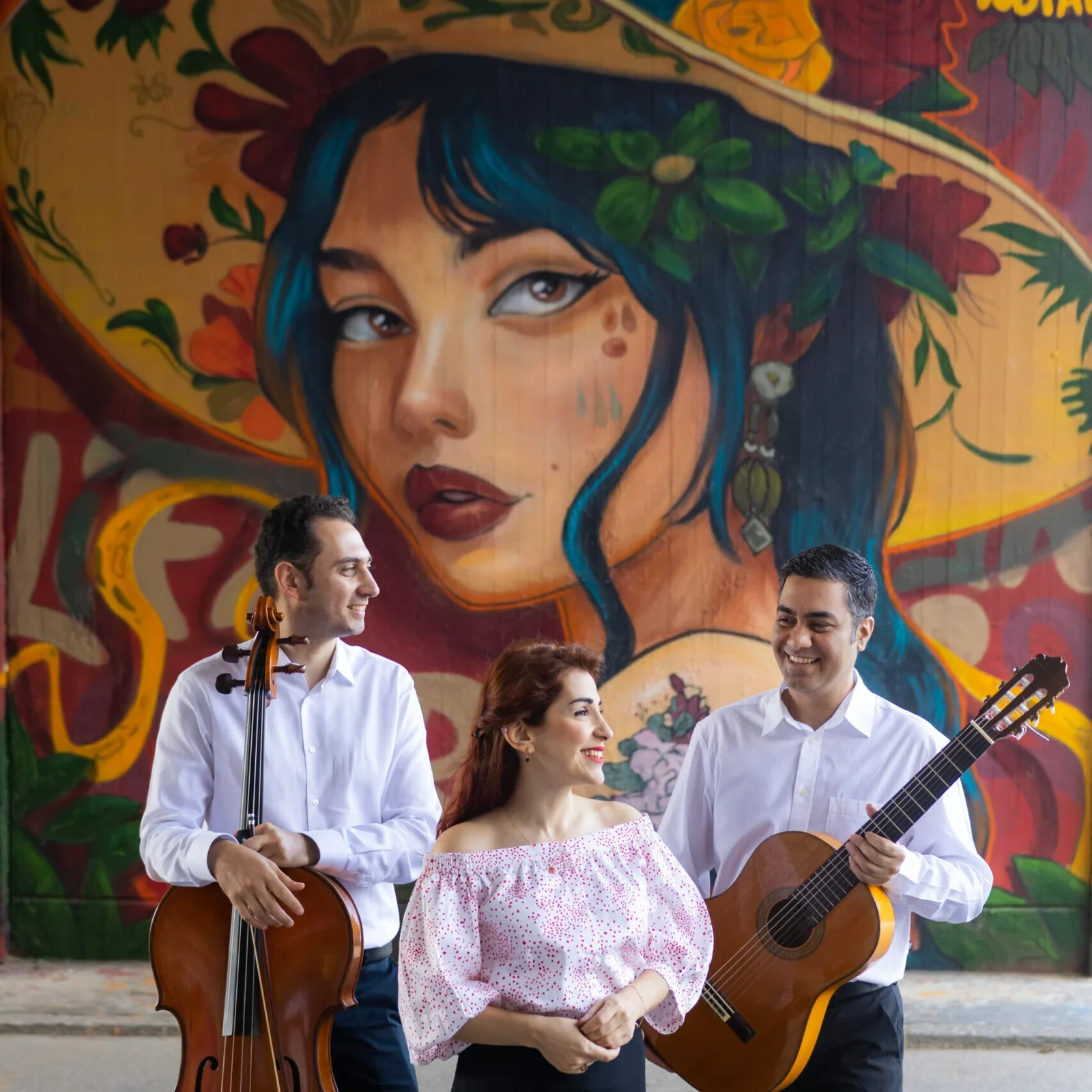 Three people with a cello and guitar in front of a colorful mural