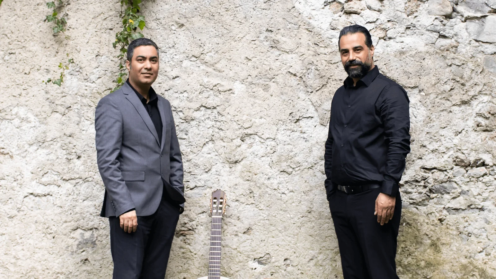 Two people standing beside an acoustic guitar against a stone wall
